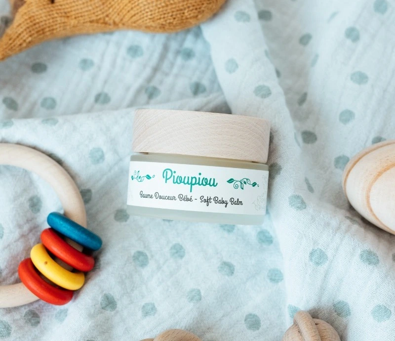 Piouspious soft baby balm jar displayed on a cozy baby blanket with wooden toys, showcasing gentle skincare for infants